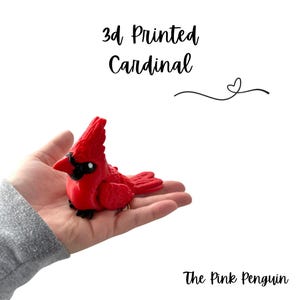 May include: A 3D-printed red cardinal figurine with black accents, held in a hand. The text "3d Printed Cardinal" is above the bird, and "The Pink Penguin" is below.