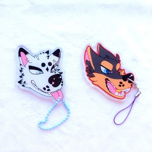 Puede incluir: Dos llaveros de cabeza de animal de dibujos animados coloridos. El primer llavero es un lobo blanco con manchas negras y una lengua rosa. El segundo llavero es un perro marrón y negro con una lengua roja. Ambos llaveros tienen un fondo de plástico transparente.
