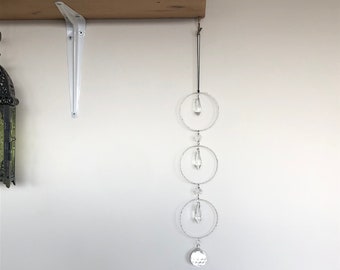 May include: A crystal suncatcher with three circular crystal prisms and a teardrop-shaped crystal hanging from a black string. The suncatcher is hanging from a white shelf bracket.