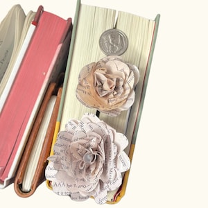 May include: Two paper flowers made from book pages, one beige and one white, are attached to a stack of books. The flowers are layered with pages from different books, creating a unique and whimsical design. The flowers are accented with a silver coin.