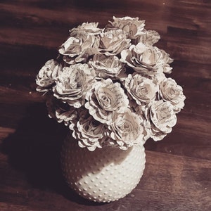 May include: A bouquet of paper roses made from old book pages in a white ceramic vase with a dotted pattern.