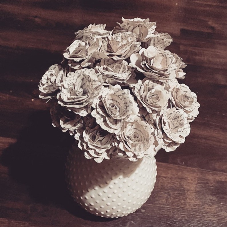 May include: A bouquet of paper roses made from old book pages in a white ceramic vase with a dotted pattern.