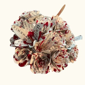 May include: A unique bouquet made of paper, featuring roses and other flower shapes. The paper is from a book, with visible text and red paint splatters. The word "DAWN" is visible on one of the petals.