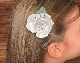 May include: A white fabric flower hair clip with sheet music print and a green leaf accent. The flower is attached to a silver metal clip and is worn in the hair.