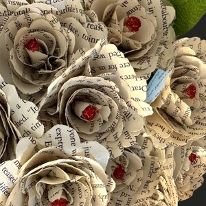 May include: A bouquet of paper roses made from old book pages. The roses are cream colored and have red glitter accents.