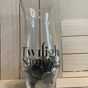 May include: A clear glass vase with the words "Twilight Saga" and "By Stephenie Meyer" printed on the side. The vase is filled with small, black and silver glass stones.