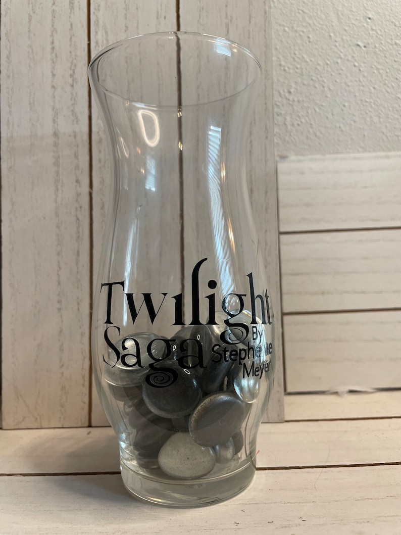 May include: A clear glass vase with the words "Twilight Saga" and "By Stephenie Meyer" printed on the side. The vase is filled with small, black and silver glass stones.