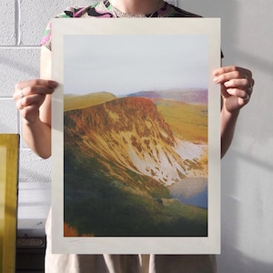 Peut inclure: Une estampe de paysage représentant une chaîne de montagnes et un lac. L'œuvre utilise des verts, des bruns et des bleus. L'estampe est tenue par une personne devant un mur de briques blanches.