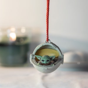 May include: A green and gray Christmas ornament featuring a character from the Star Wars franchise, The Mandalorian, in a floating pod. The ornament is hanging from a red string.