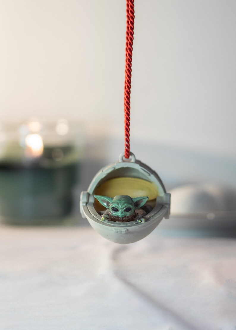 May include: A green and gray Christmas ornament featuring a character from the Star Wars franchise, The Mandalorian, in a floating pod. The ornament is hanging from a red string.