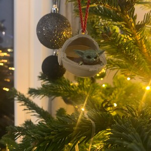 May include: A green Baby Yoda ornament hanging on a Christmas tree with lights. The ornament is in a pod-like shape and the character is looking up.