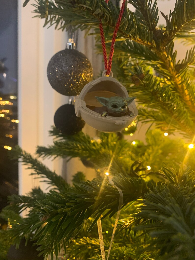 May include: A green Baby Yoda ornament hanging on a Christmas tree with lights. The ornament is in a pod-like shape and the character is looking up.