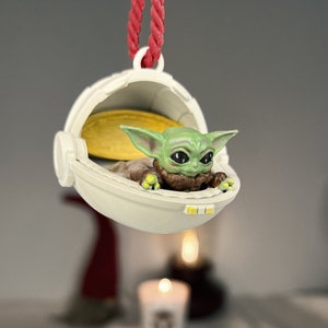 May include: A white and yellow ornament shaped like a spaceship with a small green alien figure inside. The alien has large eyes and is wearing a brown robe.