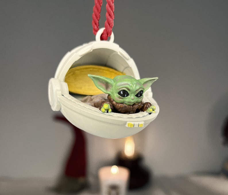 May include: A white and yellow ornament shaped like a spaceship with a small green alien figure inside. The alien has large eyes and is wearing a brown robe.