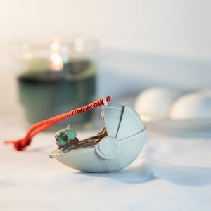 May include: A white ceramic ornament shaped like a spaceship with a small green figure inside. The ornament is attached to a red string.