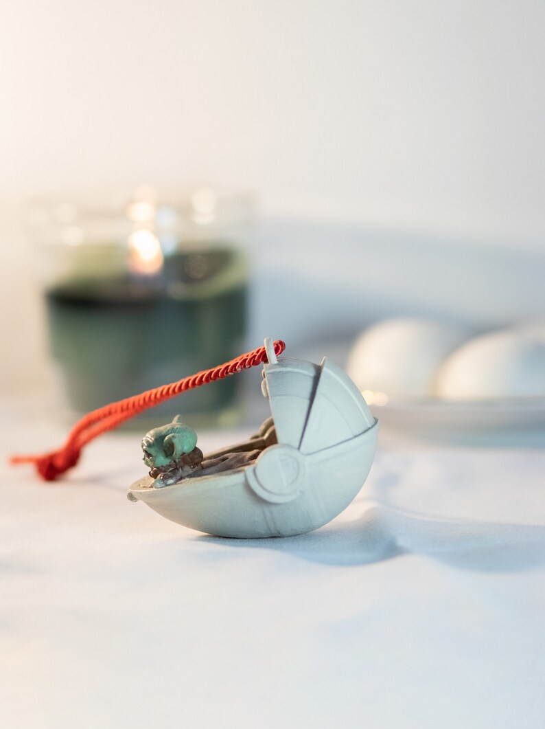May include: A white ceramic ornament shaped like a spaceship with a small green figure inside. The ornament is attached to a red string.