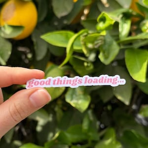 May include: A hand holding a sticker with the text "good things loading..." in pink letters. The sticker is in front of a tree with green leaves and yellow lemons.