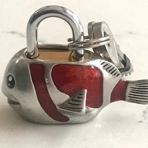 Könnte beinhalten: Ein silbernes und rotes Schloss in Fischform mit einem daran befestigten Schlüssel. Das Schloss hat eine silberne Oberfläche mit roten Emaille-Akzenten und einem detaillierten Fischdesign.