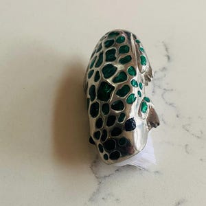May include: A silver bottle stopper with a distinctive design. It features a pattern of irregular shapes filled with green enamel, resembling leopard spots. The stopper has a white base. A decorative item.