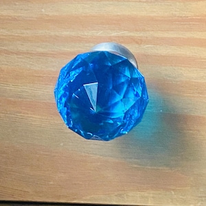 May include: A faceted, blue crystal knob with a silver base. The knob is a bright, jewel-toned blue and has a geometric, diamond-like pattern. The knob is likely for a drawer or cabinet.