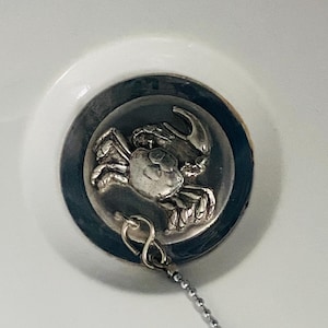 May include: A circular silver-toned sink plug featuring a raised, detailed crab emblem in the centre. The plug includes a small metal loop attached to a silver ball chain, designed to fit securely into a white basin drain opening.
