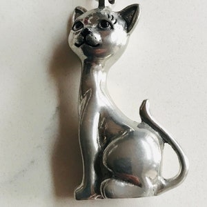 Può includere: Una statuetta di gatto color argento con occhi e ciglia neri. Il gatto è seduto con la coda arrotolata attorno al suo corpo.