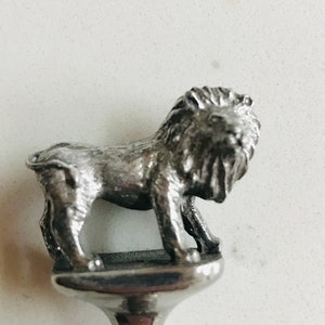 Puede incluir: Tap&oacute;n de botella de plata con un dise&ntilde;o de le&oacute;n detallado. El le&oacute;n est&aacute; de pie sobre una base con la parte superior redondeada.