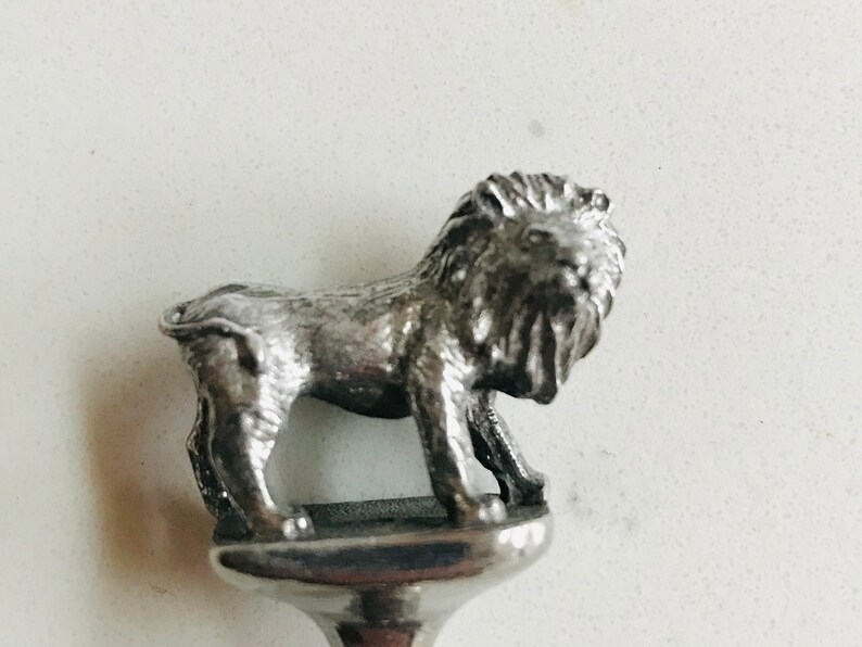 Puede incluir: Tap&oacute;n de botella de plata con un dise&ntilde;o de le&oacute;n detallado. El le&oacute;n est&aacute; de pie sobre una base con la parte superior redondeada.