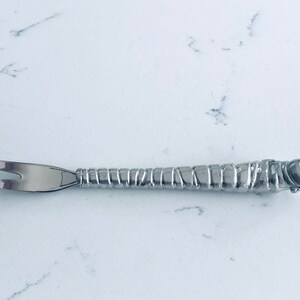May include: A silver-colored cocktail fork with a decorative handle. The handle has a textured, segmented design, and the fork has two tines. The fork is made of metal and is designed for serving appetizers or garnishes.
