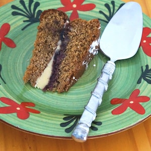 Puede incluir: Una rebanada de pastel con un relleno blanco y morado en un plato verde con flores rojas. Un servidor de pastel plateado está en el plato.