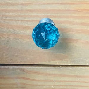 May include: A turquoise glass cabinet knob with a faceted design and a silver base. The knob is spherical and set against a light brown wooden surface. The knob's design is geometric.