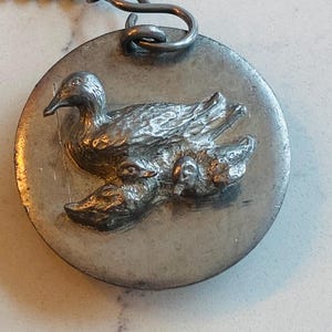 Könnte beinhalten: Ein runder, silberfarbener Anhänger mit einem detaillierten Relief einer Entenmutter und zwei Entenküken. Der Anhänger hat eine strukturierte Oberfläche und eine Metallschlaufe zum Aufhängen. Die Entenfamilie ist der Mittelpunkt.