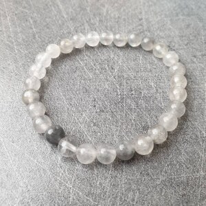 Könnte beinhalten: Ein Armband aus klarem Bergkristall mit grauen Perlen. Das Armband besteht aus runden Perlen und ist auf einem Gummiband gefädelt.