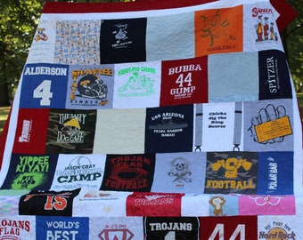 May include: A red, white, and blue patchwork quilt made from t-shirts. The quilt features various designs and text, including "Trojans", "World's Best Brother", "Bubba 44", and "Hard Rock Cafe".