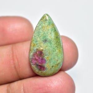 Peut inclure: Une pierre précieuse en forme de goutte d'eau, avec une base verte et des inclusions rouges vives. La pierre polie présente des inclusions rouges regroupées au centre. La pierre est tenue entre les doigts.