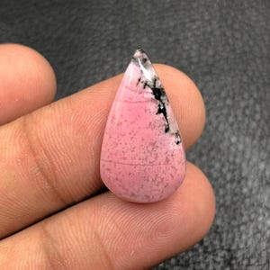 Puede incluir: Una piedra preciosa en forma de lágrima con una superficie pulida. La piedra presenta un color base rosa con patrones moteados y vetas negras y blancas cerca de la punta. La piedra preciosa se sostiene en una mano.