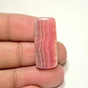 May include: A pink and white striped rectangular cabochon gemstone. The stone has a smooth, polished surface.