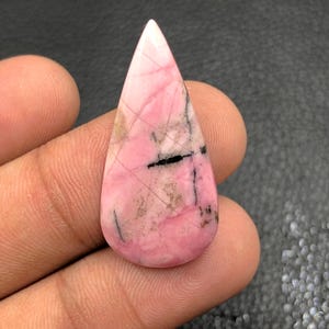 Puede incluir: Una gema pulida en forma de lágrima. La piedra presenta una mezcla de tonos rosados, con vetas más oscuras e inclusiones negras. La gema se sostiene en una mano, mostrando su tamaño y forma. Una hermosa pieza de joyería natural.