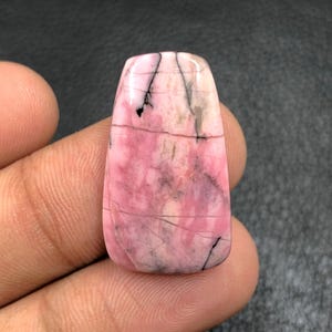 Puede incluir: Una gema rectangular pulida con bordes redondeados. La piedra presenta una mezcla de tonos rosa y blanco, con vetas grises oscuras y negras. La gema se sostiene en una mano sobre un fondo oscuro.