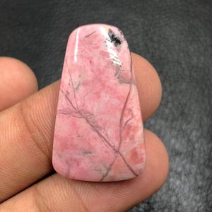 Puede incluir: Una piedra preciosa rosa pulida con forma rectangular y parte superior redondeada. La piedra presenta vetas grises oscuras y negras en toda su superficie. La piedra preciosa se sostiene en una mano, mostrando su color y textura.