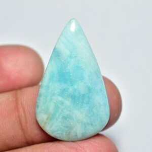 May include: A teardrop-shaped gemstone with a light blue hue and a polished surface. The stone has a marbled appearance with subtle white veining throughout. The gem is held in a person's hand.