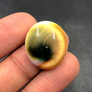 Pode incluir: Uma gema lisa, ovalada, com um padrão de redemoinho de cores amarelas, marrons e verdes. O centro da pedra tem uma área circular escura, quase preta.