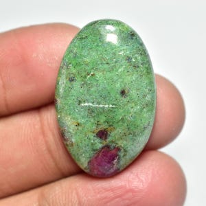 Peut inclure: Pierre précieuse de zoisite ovale, de couleur verte dominante, parsemée d'inclusions noires et rouges. La surface polie reflète la lumière, révélant les motifs et textures naturels de la pierre.