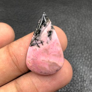 Puede incluir: Una gema en forma de lágrima con una superficie pulida. La piedra presenta una mezcla de tonos rosa, negro y gris, con una base rosa prominente y vetas negras cerca de la punta. La gema se sostiene en una mano.