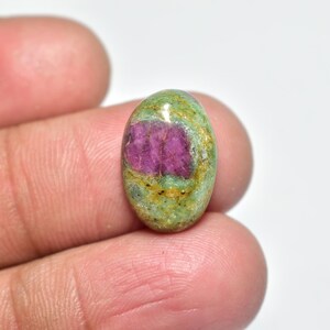 Peut inclure: Pierre précieuse ovale avec un centre fuchsia vif et un extérieur vert tacheté. La surface polie révèle un aspect naturel et terreux. La pierre est tenue entre les doigts.