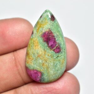 Peut inclure: Une pierre précieuse en forme de goutte d'eau, avec une base verte et des inclusions magenta vives. La pierre polie présente un mélange de teintes vertes, magenta et jaunes. Le bijou est tenu dans une main.