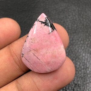 Puede incluir: Una gema pulida en forma de lágrima, de color rosa con inclusiones negras y grises. La piedra tiene una superficie lisa con vetas visibles y un acabado brillante. La gema se sostiene en una mano.