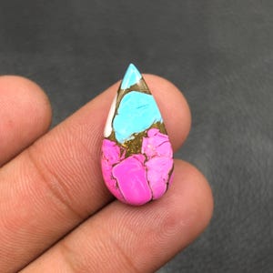 Può includere: Una gemma a forma di goccia con una vibrante tavolozza di colori. La pietra presenta una combinazione di inclusioni rosa brillante, turchese, bianco e dorato. La superficie lucida della pietra riflette la luce, evidenziando il suo motivo e la sua consistenza unici.