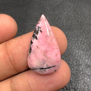 Puede incluir: Una gema pulida en forma de lágrima, con un color base rosa y vetas negras. La piedra tiene una superficie lisa y brillante y se sostiene en una mano sobre un fondo oscuro. La gema es probablemente un cabujón, adecuado para la fabricación de joyas.