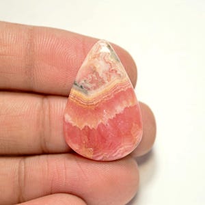 Puede incluir: Un cabujón de piedra preciosa rosa y blanca en forma de lágrima con un patrón en capas. La piedra es translúcida y tiene una superficie lisa y pulida.